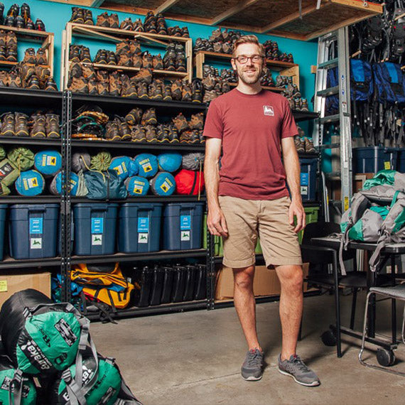 An OEN representative stands inside a gear library.  Shelves of hiking boots, tents, and sleeping bags are behind him.