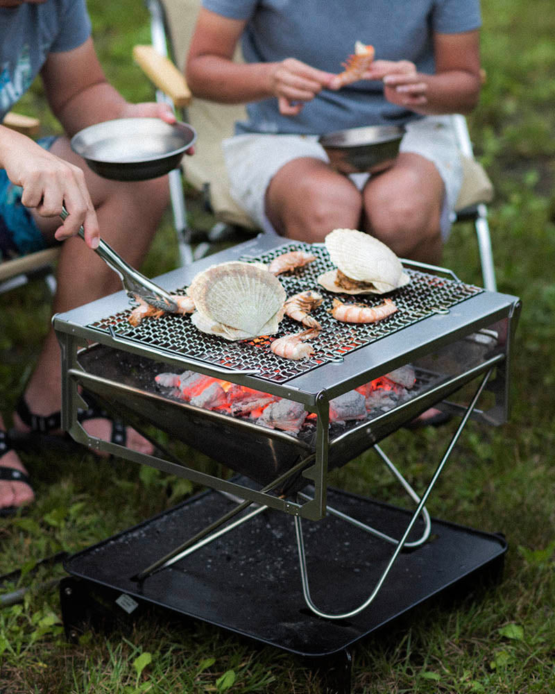 A portable camp cooking kit is being used outdoors to grill seafood. A person is using tongs to flip a scallop on the grill, and other people are sitting nearby enjoying their food.