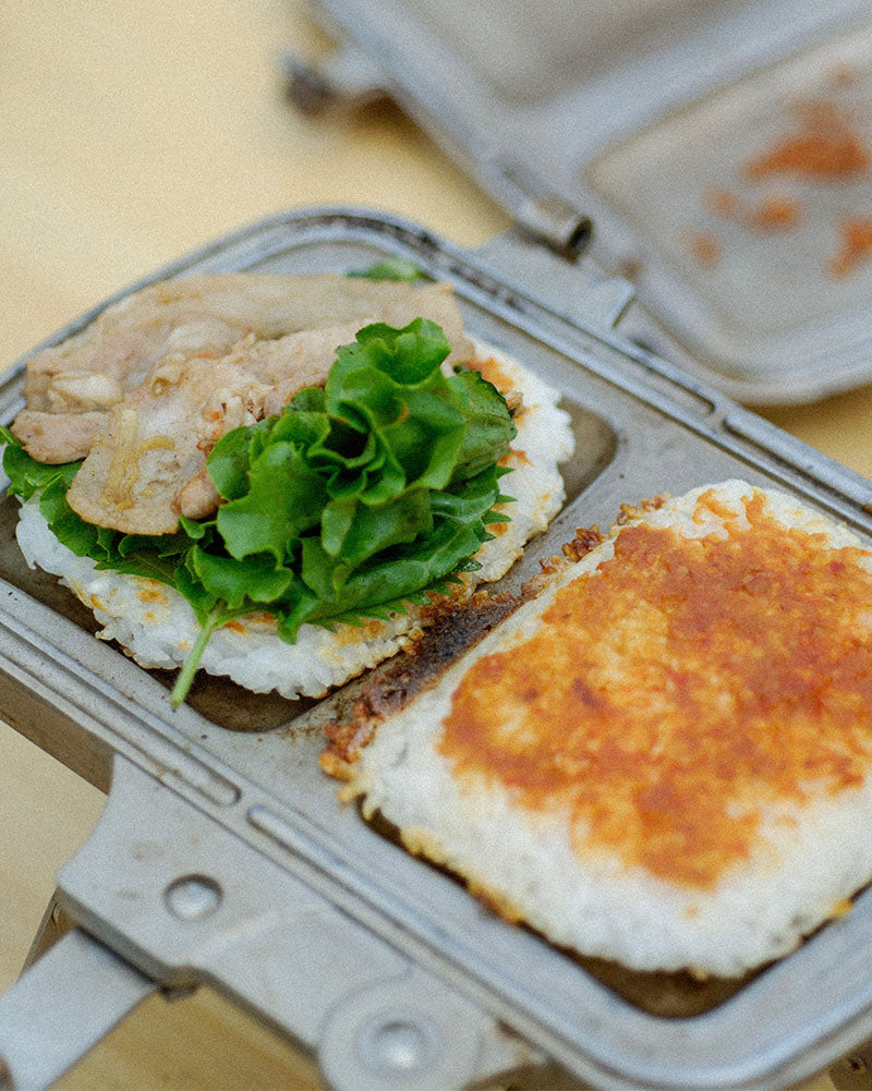 A camp cooking kit is used to prepare a sandwich on a portable camping stove. The sandwich contains meat, lettuce, and cheese between two slices of bread.