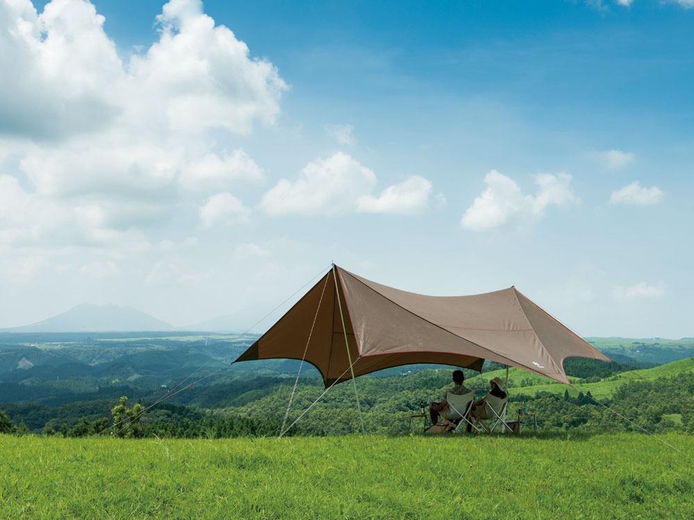 A large tarp is set up as a shelter over a camping area. Two people are sitting under the tarp enjoying the view of a mountain landscape.