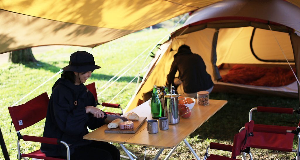 A camper prepares food at a table set up under tarp tents near a cozy, open tent in a grassy campsite.