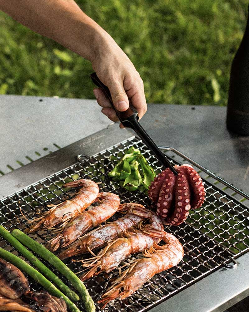A person uses tongs from their camp cooking kit to flip an octopus on a camping grill. Other food items like shrimp and asparagus are also cooked on the grill.