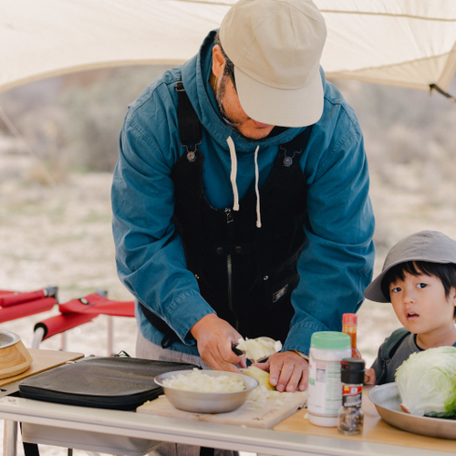 A father demonstrates how to cook on his IGT Camp Kitchen while his small child looks on.