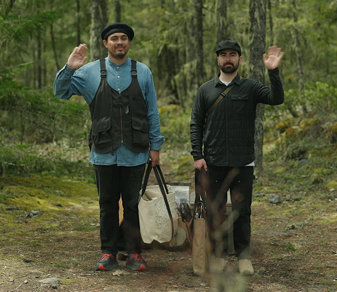 Two Snow Peak employees standing in the forest, waving, and carrying gear needed to set up a campfire.
