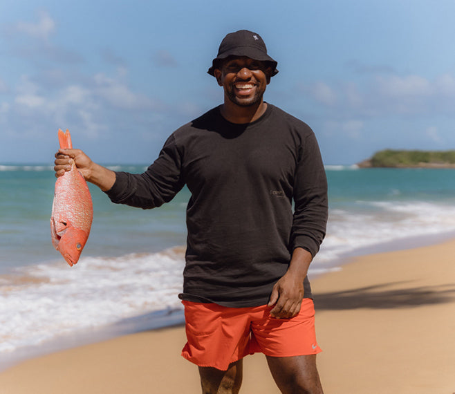 Rashad Frazier, of Camp Yoshi, stands in front of the ocean at the beach smiling and holding a bright red fish.