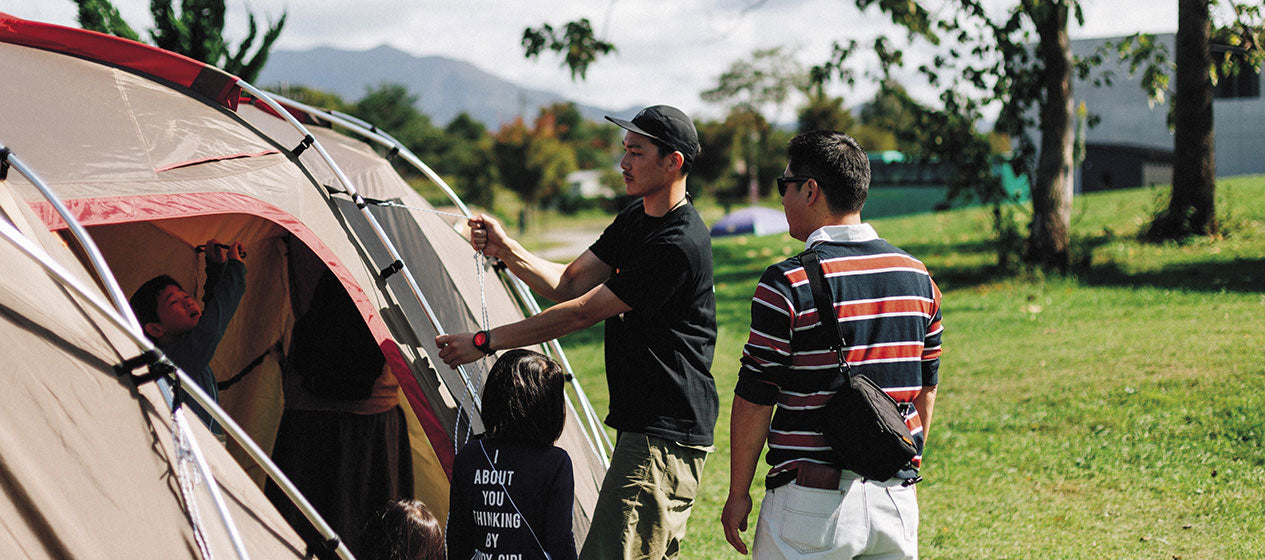 Group of campers setting up a tent at a campsite with trees in the background, preparing to use their camp cooking kit for an outdoor meal.