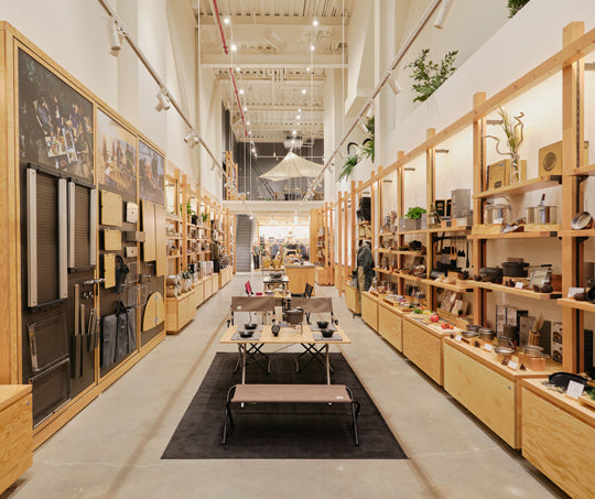 Looking into the new Snow Peak Brooklyn store, a long hall lined with light wood shelves filled with gear. Table displays run down the middle of the hall with walkways on either side.